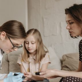 A pediatrician checks a child's blood sugar levels while her mother assists at home.